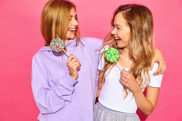 Zwei junge schöne lächelnde blonde Hippie-Frauen in der modischen Sommerkleidung. Sorglose heiße Frauen, die nahe rosa Wand aufwerfen. Positive lustige Modelle mit Lutscher