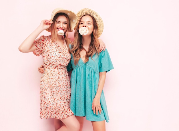 Zwei junge, schöne, lächelnde, brünette Hipster-Frauen in trendigen Sommerkleidern. Sexy sorglose Frauen, die sich in der Nähe einer rosafarbenen Wand posieren. Positive Modelle, die Blumen im Mund halten. Fröhlich und glücklich