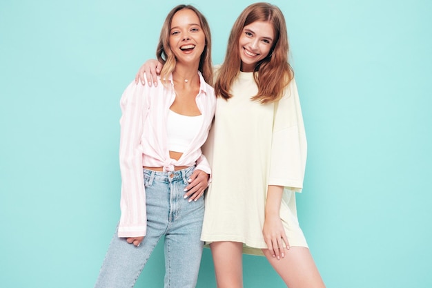 Zwei junge, schöne, lächelnde, brünette Hipster-Frauen in trendiger Sommerkleidung. Sexy sorglose Frauen, die sich in der Nähe der blauen Wand posieren. Positive Modelle, die Spaß haben Fröhlich und glücklich