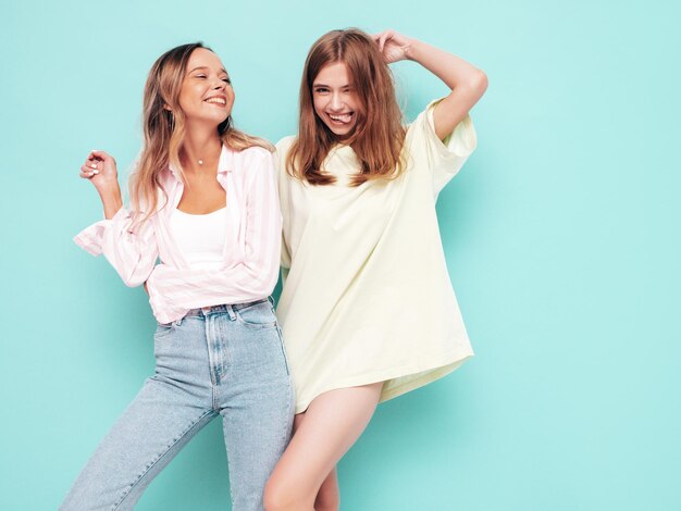 Zwei junge, schöne, lächelnde, brünette Hipster-Frauen in trendiger Sommerkleidung. Sexy sorglose Frauen, die sich in der Nähe der blauen Wand posieren. Positive Modelle, die Spaß haben Fröhlich und glücklich