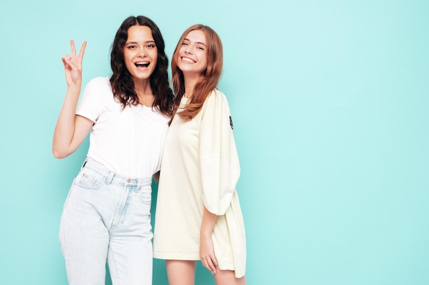 Zwei junge, schöne, lächelnde, brünette Hipster-Frauen in trendiger Sommerkleidung, sorglose Frauen, die in der Nähe der blauen Wand posieren. Positive Modelle, die Spaß haben Fröhlich und glücklich