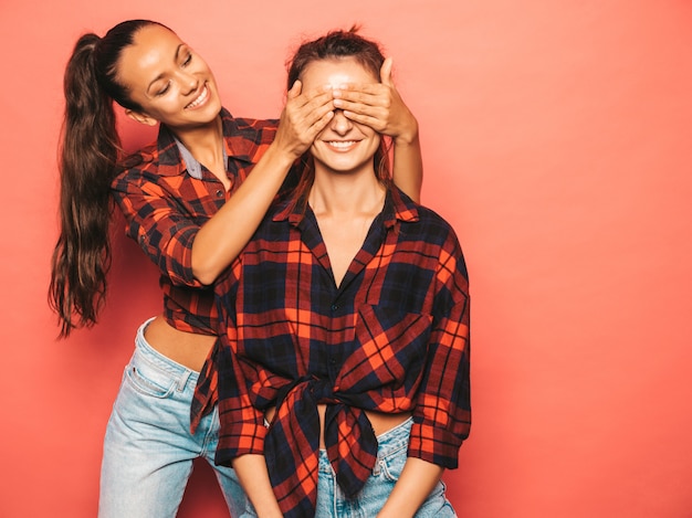 Zwei junge schöne lächelnde Brunettehippie-Mädchen in der modischen ähnlichen karierten Hemd- und Jeanskleidung. Sexy sorglose Frauen, die nahe blauer Wand im Studio aufwerfen. Abdeckung ihrer Freundaugen und -umarmen