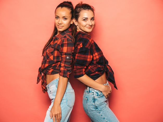 Zwei junge schöne lächelnde Brunettehippie-Mädchen in der modischen ähnlichen karierten Hemd- und Jeanskleidung Sexy sorglose Frauen, die nahe rosa Wand im Studio aufwerfen Positive Modelle, die Spaß haben