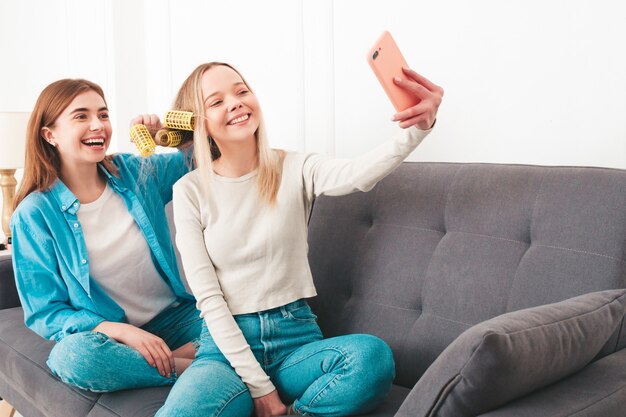 Zwei junge schöne lächelnde Frauen, die am Sofa sitzen. sorglose Modelle posieren drinnen in einer noblen Wohnung oder einem Hotelzimmer