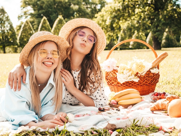 Zwei junge schöne lächelnde Frauen in trendigem Sommerkleid und Hüten. Sorglose Frauen, die draußen Picknick machen.