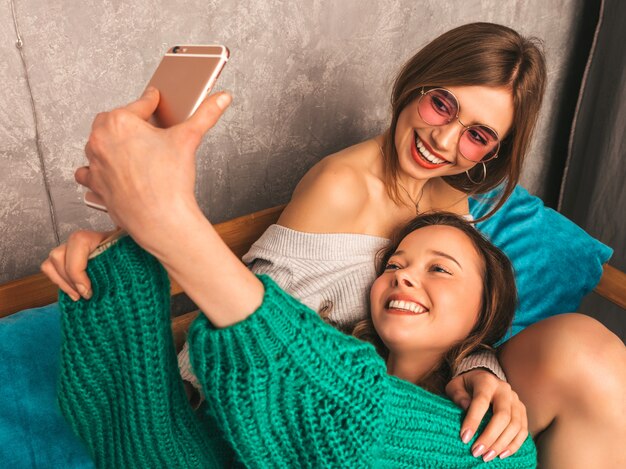 Zwei junge schöne lächelnde herrliche Mädchen in der modischen Sommerkleidung. Sexy sorglose Frauen, die im Innenraum aufwerfen und selfie nehmen. Positive Modelle, die Spaß mit Smartphone haben.