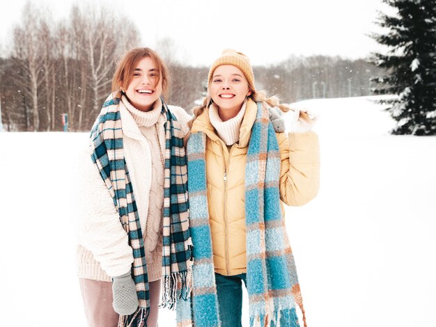 Zwei junge schöne lächelnde Hipster-Frau in trendiger warmer Kleidung und Schals. Sorglose Frauen, die auf der Straße im Park posieren. Positive pure Models, die Spaß im Schnee haben. Wintermomente genießen. Weihnachtskonzept