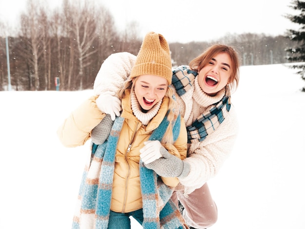 Zwei junge schöne lächelnde Hipster-Frau in trendiger warmer Kleidung und Schals. Sorglose Frauen, die auf der Straße im Park posieren. Positive pure Models, die Spaß im Schnee haben. Wintermomente genießen. Weihnachtskonzept