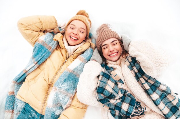 Zwei junge schöne lächelnde Hipster-Frau in trendiger warmer Kleidung und Schals. Sorglose Frauen, die auf der Straße im Park posieren. Positive reine Modelle, die im Schnee liegen. Wintermomente genießen. Weihnachtskonzept