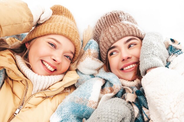 Zwei junge schöne lächelnde Hipster-Frau in trendiger warmer Kleidung und Schals. Sorglose Frauen, die auf der Straße im Park posieren. Positive reine Modelle, die im Schnee liegen. Wintermomente genießen. Weihnachtskonzept