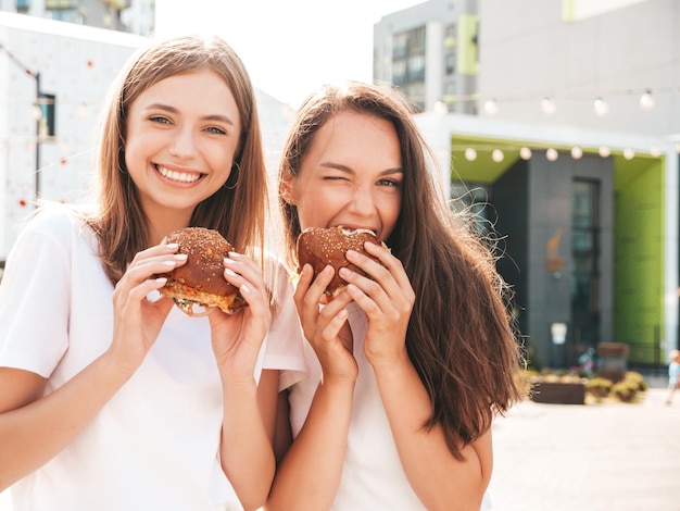 Kostenloses Foto zwei junge schöne lächelnde hipster-frauen im trendigen sommer gleiche kleidung sexy sorglose frauen posieren auf der straßepositive models, die spaß mit sonnenbrillen habenholding saftiger burger und hamburger essen