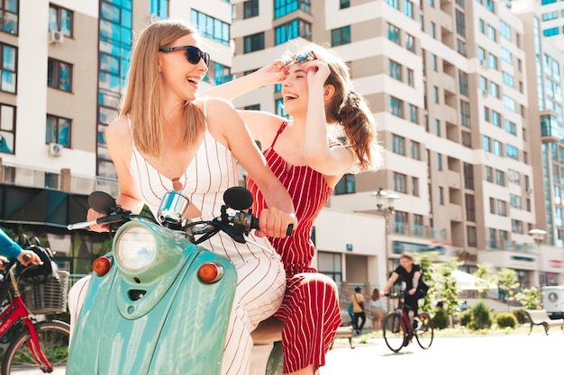 Zwei junge schöne lächelnde Hipster-Frauen in trendigen OverallsSexy sorglose Frauen, die Retro-Motorrad auf dem Straßenhintergrund fahrenPositive Models, die Spaß am klassischen italienischen Roller in Brillen haben