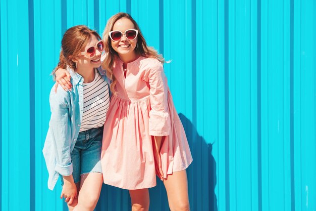 Zwei junge schöne lächelnde Hipster-Frauen in trendiger Sommerkleidung und KleidSexy sorglose Frauen posieren auf der Straße in der Nähe der blauen WandPositive reine Models, die sich bei Sonnenuntergang amüsieren