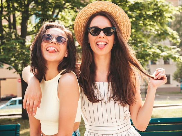 Zwei junge schöne lächelnde Hipster-Frauen in trendiger SommerkleidungSexy sorglose Frauen posieren auf dem Straßenhintergrund mit Hut Positive reine Modelle, die sich bei Sonnenuntergang amüsieren, umarmen und Zungen zeigen