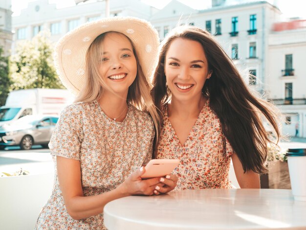 Zwei junge schöne lächelnde Hipster-Frauen in trendiger SommerkleidungSorglose Frauen, die im Veranda-Café chatten und Tee oder Kaffee trinkenPositives Modell, das Spaß hat und kommuniziert Fröhlich und glücklich