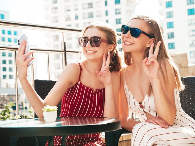 Zwei junge schöne lächelnde Hipster-Frauen in trendiger SommerkleidungSorglose Frauen posieren im Veranda-Café auf der StraßePositive Models, die Spaß haben und Selfie am Smartphone machen