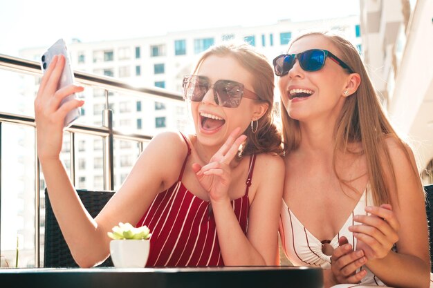 Zwei junge schöne lächelnde Hipster-Frauen in trendiger SommerkleidungSorglose Frauen posieren im Veranda-Café auf der StraßePositive Models, die Spaß haben und Selfie am Smartphone machen