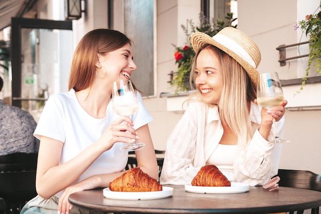 Zwei junge schöne lächelnde Hipster-Frauen in trendiger SommerkleidungSorglose Frauen posieren im Veranda-Café auf der StraßePositive Models trinken WeißweinGenießen ihren UrlaubCroissant essen