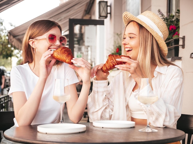 Zwei junge schöne lächelnde Hipster-Frauen in trendiger SommerkleidungSorglose Frauen posieren im Veranda-Café auf der StraßePositive Models trinken WeißweinGenießen ihren UrlaubCroissant essen