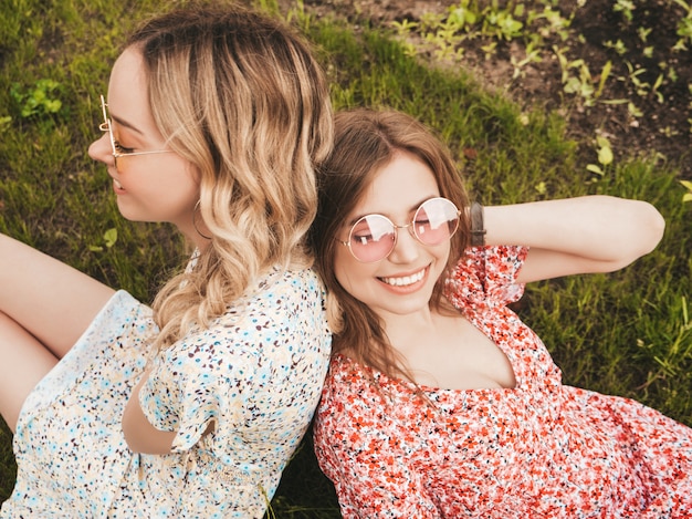Kostenloses Foto zwei junge schöne lächelnde hipster-mädchen im trendigen sommer-sommerkleid. sexy sorglose frauen, die auf dem grünen gras in der sonnenbrille liegen. positive modelle, die spaß haben