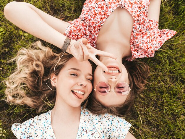 Zwei junge schöne lächelnde Hipster-Mädchen im trendigen Sommer-Sommerkleid. Sexy sorglose Frauen, die auf dem grünen Gras in der Sonnenbrille liegen. Positive Modelle, die Spaß haben