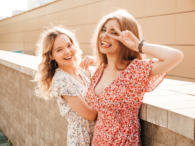 Zwei junge schöne lächelnde Hipster-Mädchen im trendigen Sommer-Sommerkleid. Sexy sorglose Frauen, die auf dem Straßenhintergrund aufwerfen. Positive Modelle, die Spaß haben und Friedenszeichen zeigen