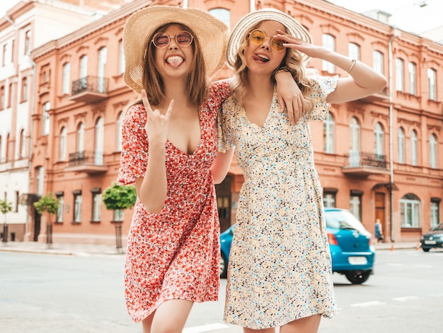 Zwei junge schöne lächelnde hipster-mädchen im trendigen sommer-sommerkleid. sexy sorglose frauen, die auf dem straßenhintergrund in den hüten aufwerfen. positive models, die spaß haben und sich umarmen