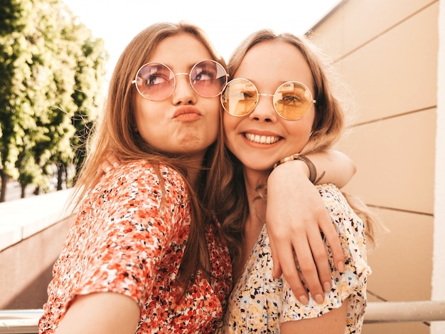 Zwei junge schöne lächelnde Hipster-Mädchen im trendigen Sommer-Sommerkleid. Sexy sorglose Frauen, die auf dem Straßenhintergrund in der Sonnenbrille aufwerfen. Sie machen Selfie-Selbstporträtfotos auf dem Smartphone bei Sonnenuntergang