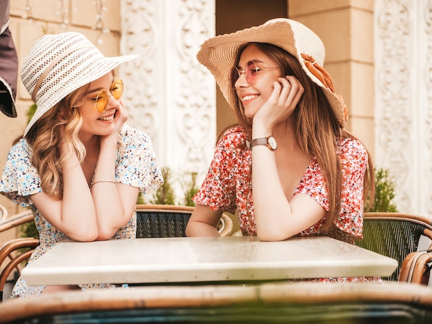 Zwei junge schöne lächelnde Hipster-Mädchen im trendigen Sommer-Sommerkleid. Sorglose Frauen, die im Veranda-Café auf dem Straßenhintergrund in der Sonnenbrille plaudern. Positive Modelle, die Spaß haben und kommunizieren