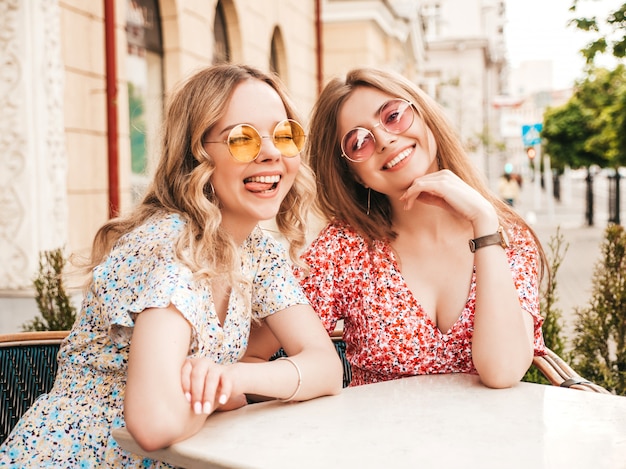 Zwei junge schöne lächelnde Hipster-Mädchen im trendigen Sommer-Sommerkleid. Sorglose Frauen, die im Veranda-Café auf dem Straßenhintergrund in der Sonnenbrille plaudern. Positive Modelle, die Spaß haben und kommunizieren