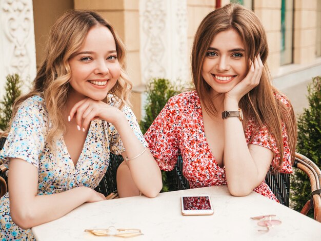 Zwei junge schöne lächelnde Hipster-Mädchen im trendigen Sommer-Sommerkleid. Sorglose Frauen, die im Veranda-Café auf dem Straßenhintergrund plaudern. Positive Modelle, die Spaß haben und kommunizieren
