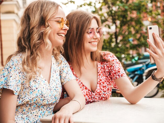 Zwei junge schöne lächelnde Hipster-Mädchen im trendigen Sommer-Sommerkleid. Sorglose Frauen, die im Veranda-Café auf dem Straßenhintergrund plaudern. Positive Modelle, die Spaß haben und Selfie auf Smartphone nehmen