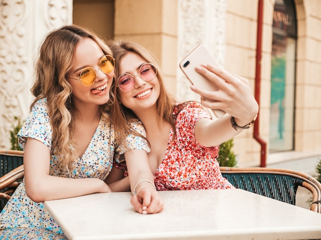 Zwei junge schöne lächelnde Hipster-Mädchen im trendigen Sommer-Sommerkleid. Sorglose Frauen, die im Veranda-Café auf dem Straßenhintergrund plaudern. Positive Modelle, die Spaß haben und Selfie auf Smartphone nehmen