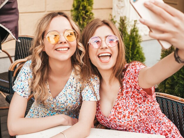 Zwei junge schöne lächelnde Hipster-Mädchen im trendigen Sommer-Sommerkleid. Sorglose Frauen, die im Veranda-Café auf dem Straßenhintergrund plaudern. Positive Modelle, die Spaß haben und Selfie auf Smartphone nehmen