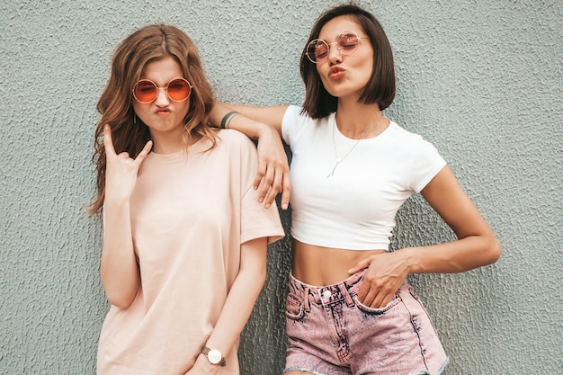 Zwei junge schöne lächelnde Hipster-Mädchen in der trendigen Sommerkleidung. Sexy sorglose Frauen, die auf der Straße nahe Wand in der Sonnenbrille aufwerfen. Positive Models, die Spaß haben und Rock'n'Roll-Zeichen zeigen
