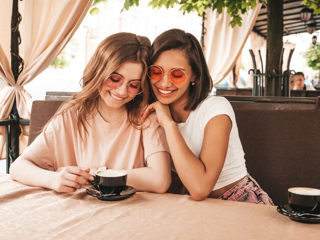 Zwei junge schöne lächelnde Hipster-Mädchen in trendigen Sommer-Freizeitkleidung. Sorglose Frauen, die im Veranda-Terrassencafé plaudern und Kaffee trinken. Positive Modelle, die Spaß haben und kommunizieren