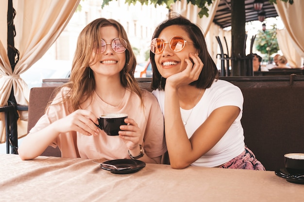 Zwei junge schöne lächelnde Hipster-Mädchen in trendigen Sommer-Freizeitkleidung. Sorglose Frauen, die im Veranda-Terrassencafé plaudern und Kaffee trinken. Positive Modelle, die Spaß haben und kommunizieren