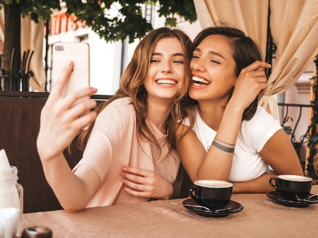 Zwei junge schöne lächelnde Hipster-Mädchen in trendigen Sommerkleidern. Sorglose Frauen, die im Veranda-Café plaudern und Kaffee trinken. Positive Modelle, die Spaß haben und Selfie auf Smartphone nehmen