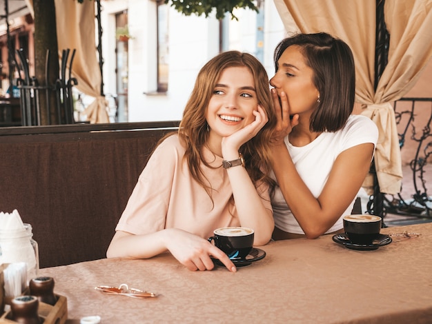 Zwei junge schöne lächelnde Hipster-Mädchen in trendiger Sommerkleidung. Sorglose Frauen, die im Veranda-Café plaudern und Kaffee trinken. Positives Modell teilt Geheimnis zu ihrem Freundohr