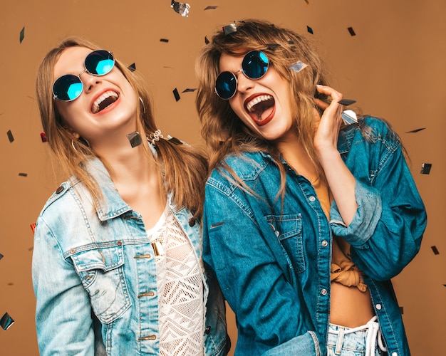 Zwei junge schöne lächelnde Mädchen in der modischen Sommerkleidung und -sonnenbrille. Sexy sorglose Frauenaufstellung. Positive schreiende Modelle unter Konfetti