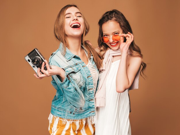 Zwei junge schöne lächelnde Mädchen in der zufälligen Kleidung und in der Sonnenbrille des modischen Sommers. Sexy sorglose Frauenaufstellung. Fotografieren mit der Retro-Kamera