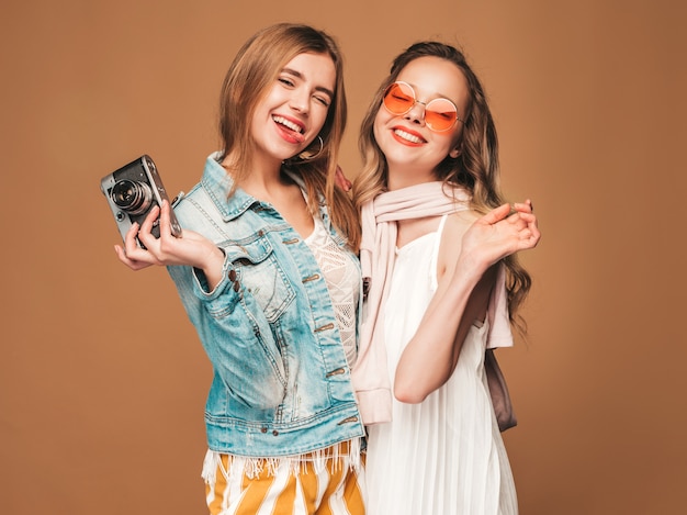 Zwei junge schöne lächelnde Mädchen in der zufälligen Kleidung und in der Sonnenbrille des modischen Sommers. Sexy sorglose Frauenaufstellung. Fotografieren mit der Retro-Kamera