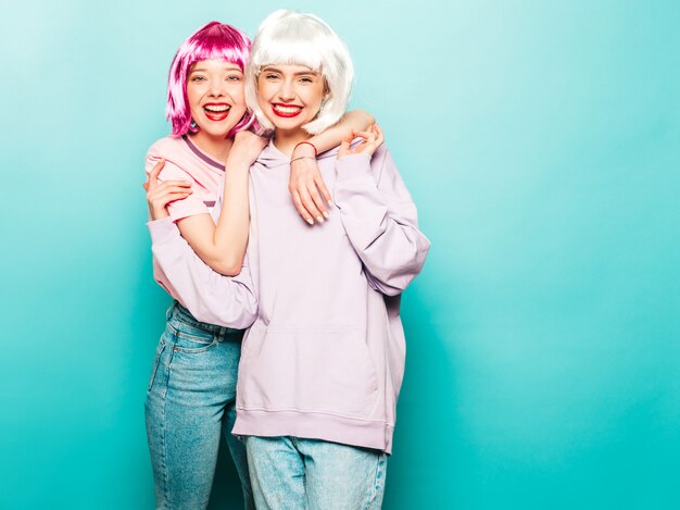 Zwei junge sexy Hipster-Mädchen in Perücken und roten Lippen. Schöne trendige Frauen in Sommerkleidung. Sorglose Modelle, die nahe blaue Wand im Studio aufstellen, das verrückt wird