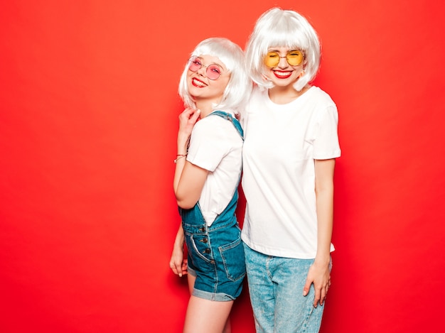 Zwei junge sexy lächelnde Hipster-Mädchen in den weißen Perücken und in den roten Lippen. Schöne trendige Frauen in der Sommerkleidung