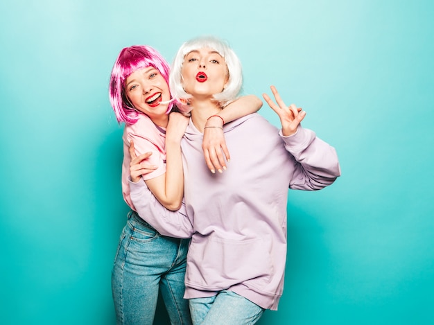 Zwei junge sexy lächelnde Hipster-Mädchen in Perücken und roten Lippen. Schöne trendige Frauen in Sommerkleidung. Sorglose Modelle, die nahe blaue Wand im Studio aufwerfen, zeigen Friedenszeichen