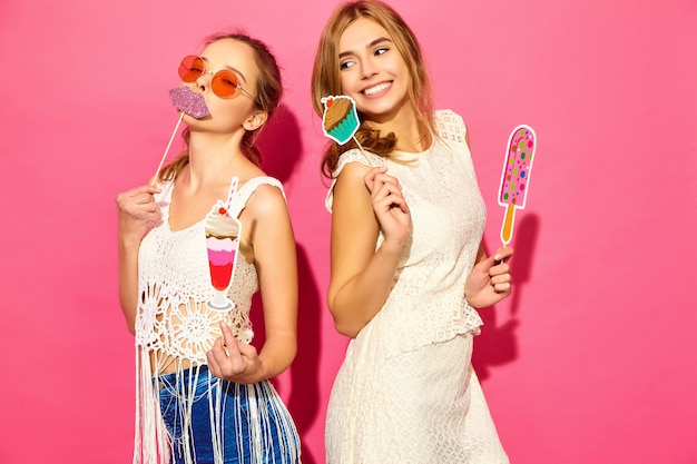 Zwei junge stilvolle lächelnde blonde Frauen, die süße Eiscreme der Stützen und gefälschtes Cocktail essen. Positive Modelle im Sommerhippie kleiden die Aufstellung nahe rosa Wand in der Sonnenbrille