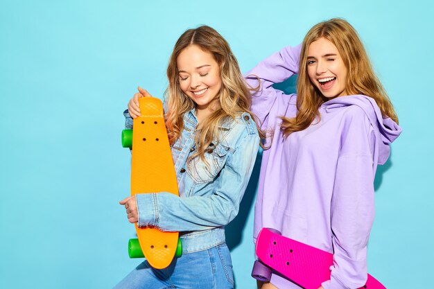 Zwei junge stilvolle lächelnde blonde Frauen mit Penny-Skateboards. Frauen in der Sommerhippie-Sportkleidung, die nahe blauer Wand aufwirft. Positive Vorbilder