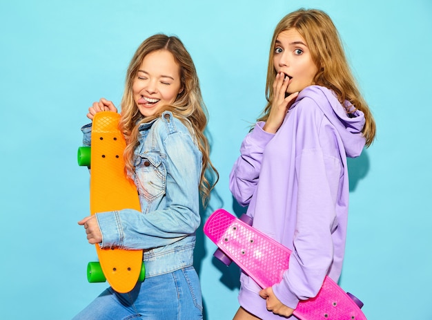 Zwei junge stilvolle lächelnde blonde Frauen mit Penny-Skateboards. Frauen in der Sommerhippie-Sportkleidung, die nahe blauer Wand aufwirft. Positive Vorbilder