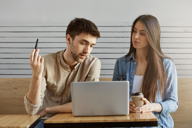 Zwei junge Studenten in stilvoller Kleidung sitzen in der Cafeteria und sprechen über das Studienprojekt und schauen auf den Laptop-Monitor, um eine Lösung zu finden