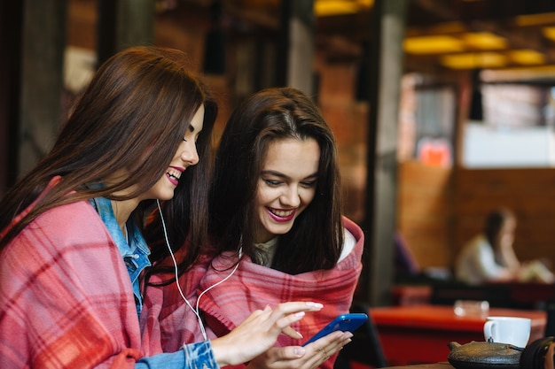Zwei junge und schöne Mädchen sitzen am Tisch und hören Musik mit einem Smartphone